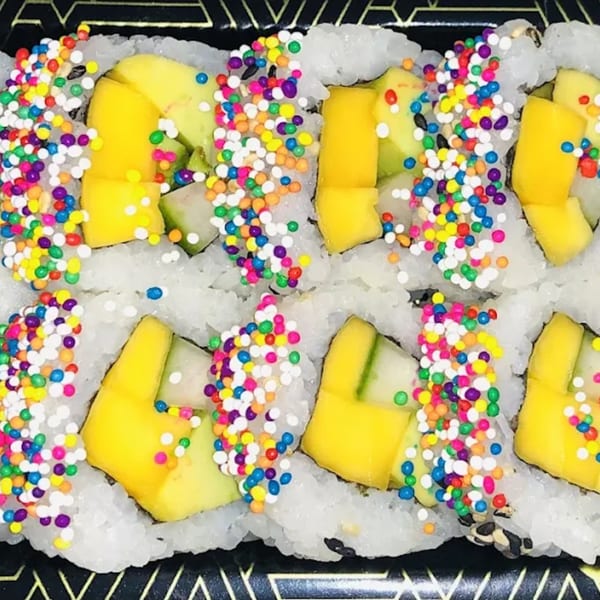 Colorful sushi rolls with mango, avocado, and rainbow sprinkles on a black tray.